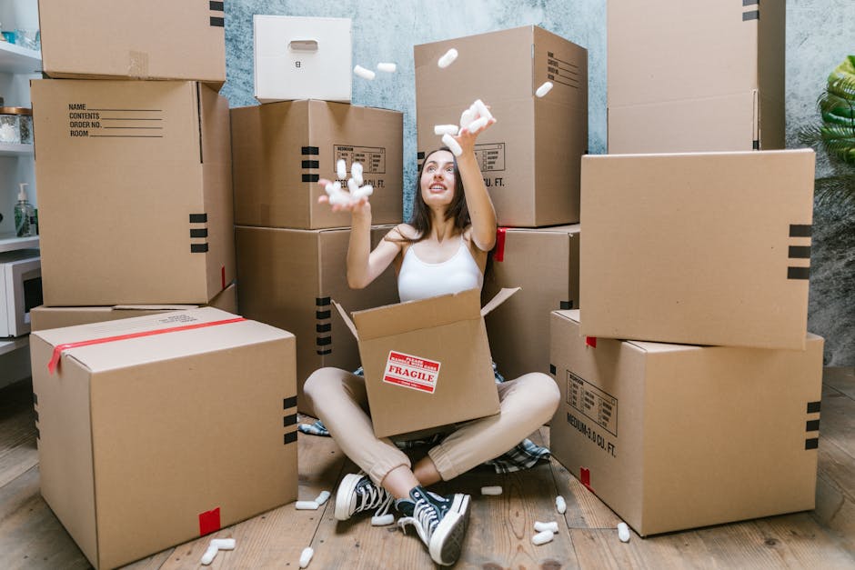 A young woman is sitting on a wooden floor surrounded by numerous large cardboard moving boxes of various sizes, some sealed with red packing tape and others open, revealing packing materials. She is inside a room with a light blue textured wall and a small potted plant visible on the right. The woman is wearing casual clothing, including a white tank top, beige pants, and striped sneakers. She is joyfully tossing packing peanuts into the air, with some falling around her. The boxes are stacked behind and around her, with labels indicating contents and fragility, suggesting an ongoing home relocation process. Nearby, a white microwave and some other household items are partially visible, and the scene conveys the active packing phase of furniture transport and house removals handled by Removal Van Haringey, emphasizing the logistics involved in home moving.