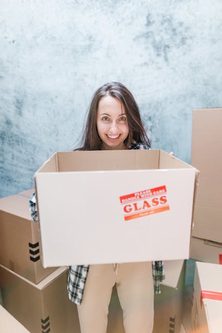 Close-up of a person wearing a dark teal shirt and navy blue pants using their right hand to hold a large corrugated cardboard box inside a property, with a red and white caution label indicating 'Cau