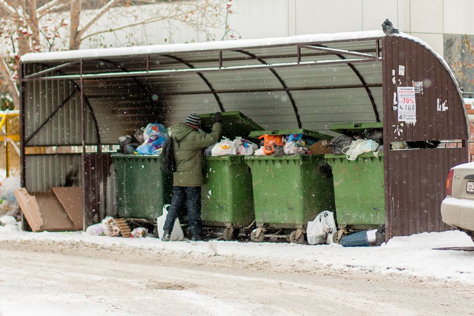 A person wearing a green coat and striped hat standing outside on a snowy pavement, collecting items from a collection of large green and brown wheelie bins situated inside a covered outdoor waste dis