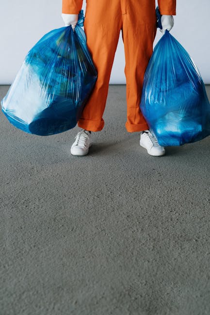 A person wearing a green coat and striped hat standing outside on a snowy pavement, collecting items from a collection of large green and brown wheelie bins situated inside a covered outdoor waste dis