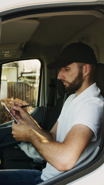 A man with a beard, wearing a black cap and white polo shirt, is seated inside a vehicle, likely a removal van. He is holding a smartphone in his right hand and using his left hand to assist with the
