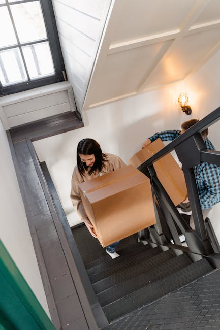 A woman with dark hair, dressed in a beige top, is carrying a large cardboard box down a narrow indoor staircase with black carpeting. Behind her, a man in a blue and black checked shirt is also holding a box, partially obscured by the staircase railing. The staircase is situated inside a well-lit residential building with white walls, a wooden window frame, and a small wall-mounted light fixture emitting warm light. The scene depicts the process of home relocation, with removal services by Removal Van Haringey, involving furniture transport and packing during a house move. The surroundings include the stairwell and doorway leading to the ground floor, with additional packing materials or boxes likely part of the ongoing moving process.