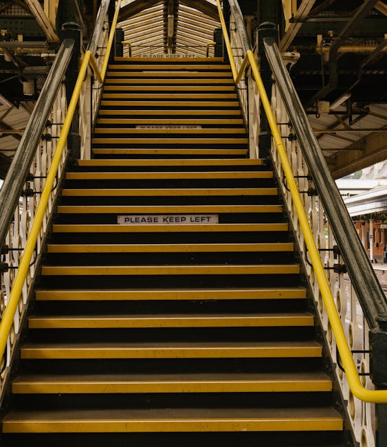 A wide, indoor staircase with yellow handrails on both sides and black and yellow striped anti-slip treads on each step, leading up to a higher level within a building. The steps are made of metal or wood, and there is a sign on one of the middle steps reading 'Please Keep Left' to guide movement during the loading or unloading process. The surrounding structure includes visible metal framework and beams, indicating a storage or loading area, typical in a house removal or logistics setting. Natural lighting illuminates the staircase, with parts of the upper floor or platform faintly visible at the top. This scene reflects the logistics involved in furniture transport and home relocation, with clean, organized staging suitable for moving operations like those performed by Removal Van Haringey.