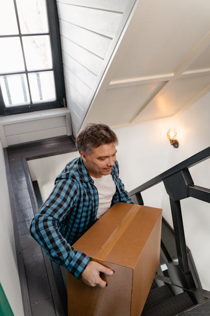 A woman with dark hair, dressed in a beige top, is carrying a large cardboard box down a narrow indoor staircase with black carpeting. Behind her, a man in a blue and black checked shirt is also holding a box, partially obscured by the staircase railing. The staircase is situated inside a well-lit residential building with white walls, a wooden window frame, and a small wall-mounted light fixture emitting warm light. The scene depicts the process of home relocation, with removal services by Removal Van Haringey, involving furniture transport and packing during a house move. The surroundings include the stairwell and doorway leading to the ground floor, with additional packing materials or boxes likely part of the ongoing moving process.