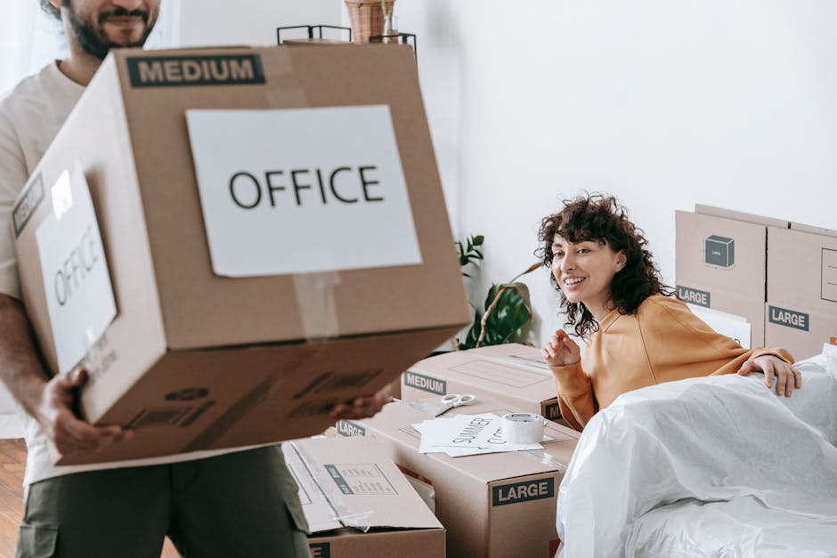 In a residential interior, a person is seen holding two large cardboard boxes labeled 'OFFICE' with one partially obscured by another box, while a woman with curly hair and a warm smile is sitting on a bed surrounded by packed moving boxes of various sizes, some labeled 'LARGE' and 'MEDIUM'. The scene depicts a home relocation process, with furniture and packing materials such as cardboard boxes, paper, and packing tape visible. The background includes a potted plant, a window with natural light, and cardboard boxes stacked on the floor and against the wall, emphasizing the packing stage of an office or household move. This setting illustrates the packing and moving logistics handled by Removal Van Haringey as part of their house and office removal services, highlighting the careful organization and preparation involved in furniture transport and home relocation within Haringey.