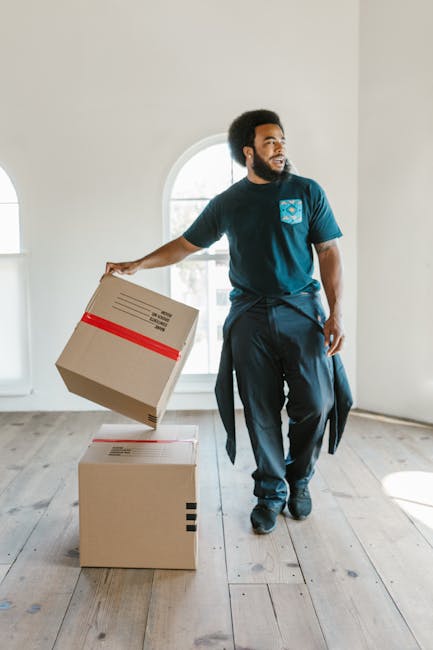 A man with a beard and dark curly hair stands inside a well-lit room with white walls and a large arched window, wearing a blue t-shirt with a logo and dark trousers. He is in the process of moving, holding a cardboard box wrapped with red tape in his right hand, preparing to place it onto a second box on the wooden floor. The second box, also cardboard, is positioned directly beneath the moving box and appears to be ready for stacking during a home relocation or furniture transport process. The room has a minimalistic appearance with no furniture visible, and natural daylight illuminates the scene. This image, associated with Removal Van Haringey's office removals service in Haringey, north Tottenham, depicts the loading or packing phase of a professional moving service, showcasing the careful handling of household or business items during a physical move.