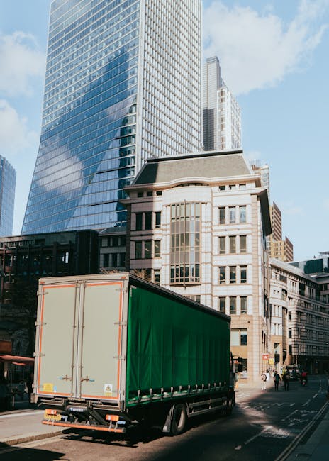 A large green removal truck parked on a city street in front of modern glass skyscrapers and older stone buildings, with the vehicle's rear doors closed and cargo area visible. The truck is positioned near a building entrance, with a pavement and road markings, and there are a few pedestrians and vehicles in the background. The scene appears to be in a busy urban area suitable for home relocation and furniture transport services provided by Removal Van Haringey, highlighting the logistics of professional removals and loading processes in a city environment.