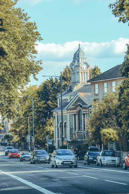 A street scene in Wood Green, N22, featuring a row of parked cars along the roadside, including sedans and hatchbacks, with some vehicles partially visible and others fully in view. Behind the cars, there are residential buildings with brick facades and pitched roofs, interspersed with lush green trees providing shade and a natural backdrop. In the background, a historic clock tower with a spire, large clock face, and ornate architectural details rises above the surrounding trees and buildings, indicating a notable local landmark. The sky is clear with a few scattered clouds, and the overall scene suggests a calm suburban area with active vehicle traffic. This setting is relevant to house removals and furniture transport, as Removal Van Haringey manages logistical moving services within such urban and residential environments, ensuring seamless loading and transportation of household items during home relocations.